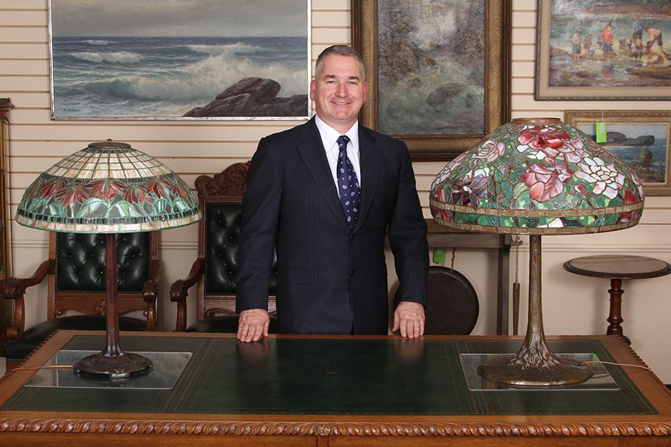 A man standing behind a desk in front of two lamps.